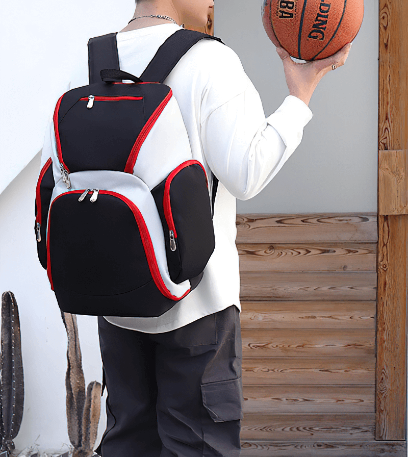 Man wearing waterproof lightweight football backpack holding basketball, featuring large capacity and sleek red-trimmed design.